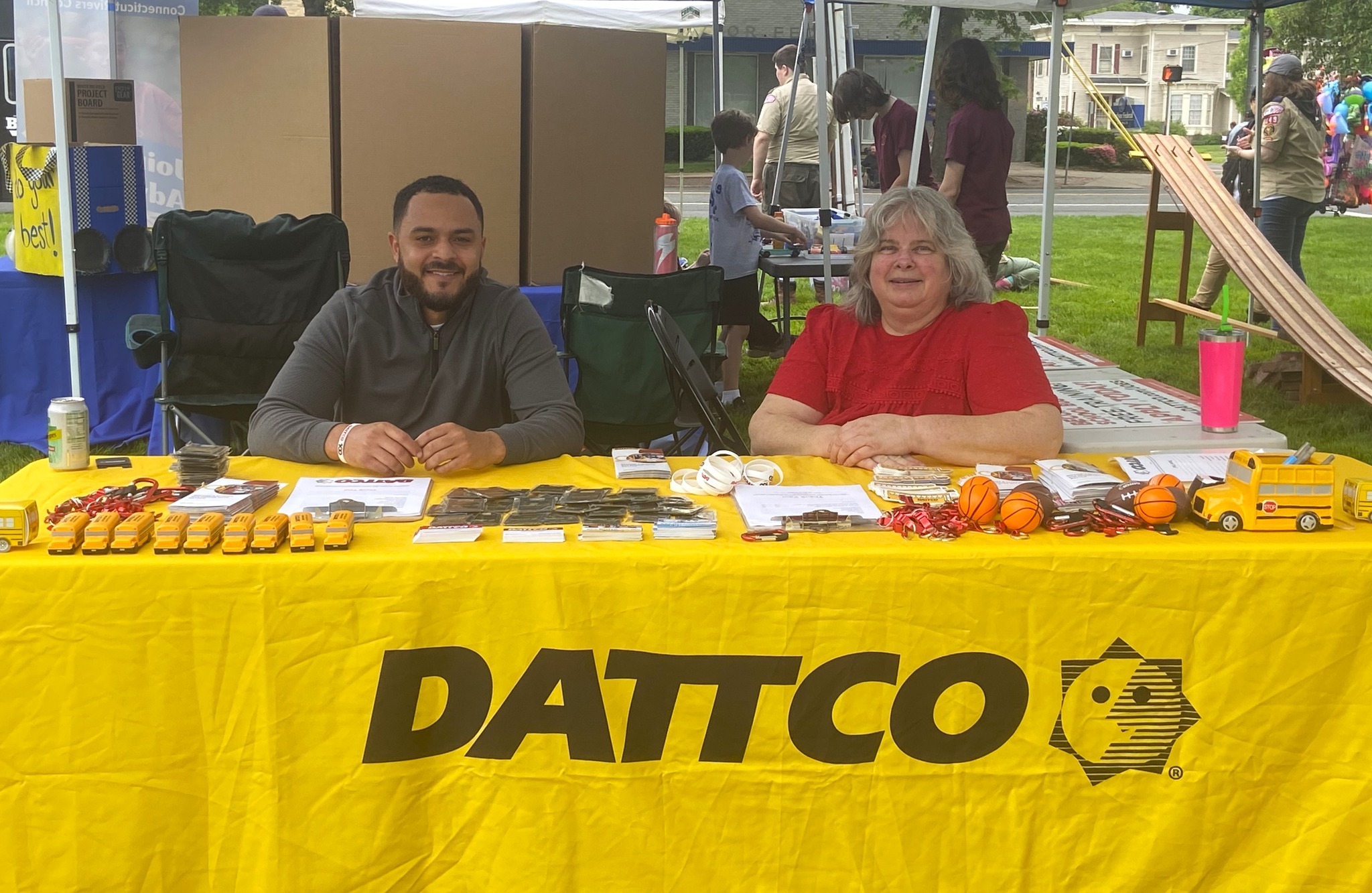 A yellow tablecloth with the logo "DATTCO" is spread out on a table at an outdoor event. Two individuals, a man and a woman, are seated behind the table. The man is wearing a gray jacket and is smiling, while the woman is in a red shirt and also smiling. Various promotional items are displayed on the table, including small yellow school bus models, keychains, and other giveaways. In the background, there are tents and people engaging in activities. A pink drink cup is visible on the table as well.