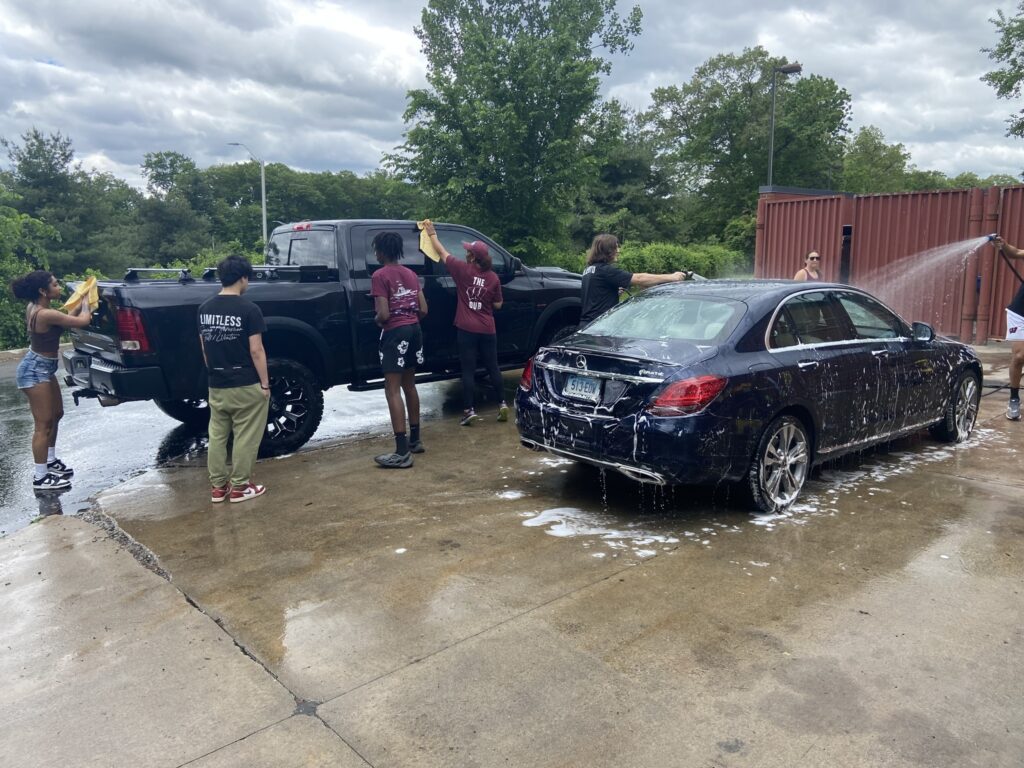 A group of individuals is washing two vehicles at a car wash. On the left, a black pickup truck is being cleaned, with two people using yellow cloths to wipe it down. On the right, a dark blue sedan is being rinsed with water, creating a foamy appearance on its surface. The scene is set outdoors, with cloudy skies and greenery in the background. The ground is wet, reflecting the vehicles and the surrounding area.