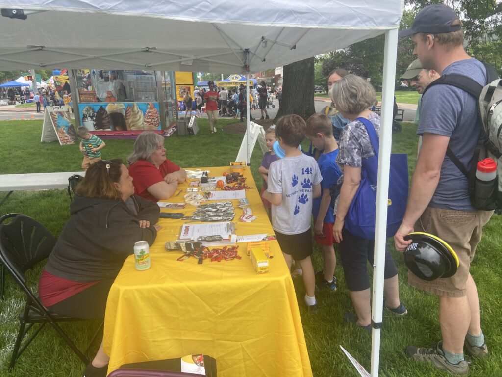 A vibrant outdoor scene at a fair features a yellow table under a white canopy. Two women sit at the table, surrounded by various items like pamphlets and small giveaways. A group of children and adults gathers around the table, engaging with the women. In the background, colorful food stalls and people enjoying the event can be seen, adding to the lively atmosphere. A child in a striped shirt plays nearby on the grass.