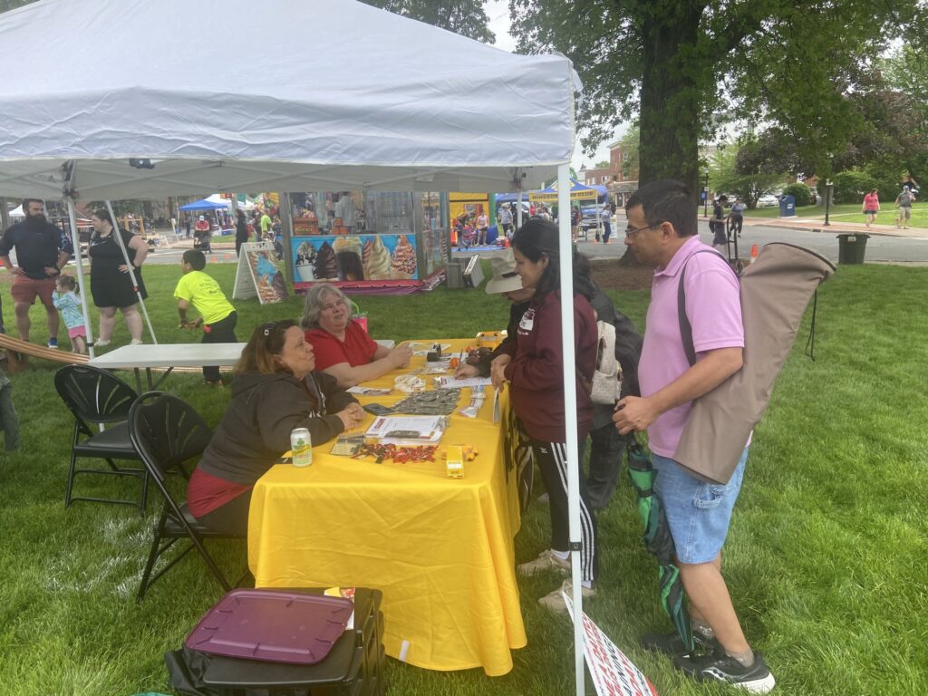 A tent with a yellow tablecloth is set up in a grassy area, where two women are seated at the table, engaged in conversation. One woman is wearing a black jacket and the other has gray hair and is dressed in red. A man in a pink shirt stands nearby, holding a large, rolled-up item, possibly an umbrella. Another woman is standing at the table, writing something down. In the background, there are people enjoying the event, with colorful booths and attractions visible.